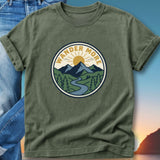 Olive green t-shirt with circular outdoor graphic: "WANDER MORE" text, mountains, sun, and river.