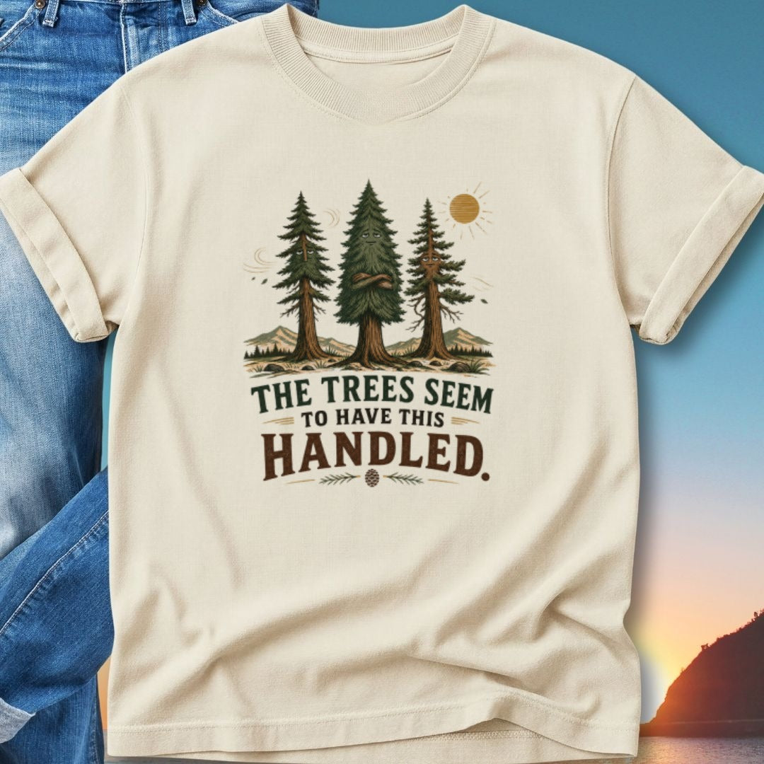 Cream t-shirt with "The Trees Seem to Have This Handled" text and pine trees graphic.