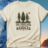 Cream t-shirt with "The Trees Seem to Have This Handled" text and pine trees graphic.