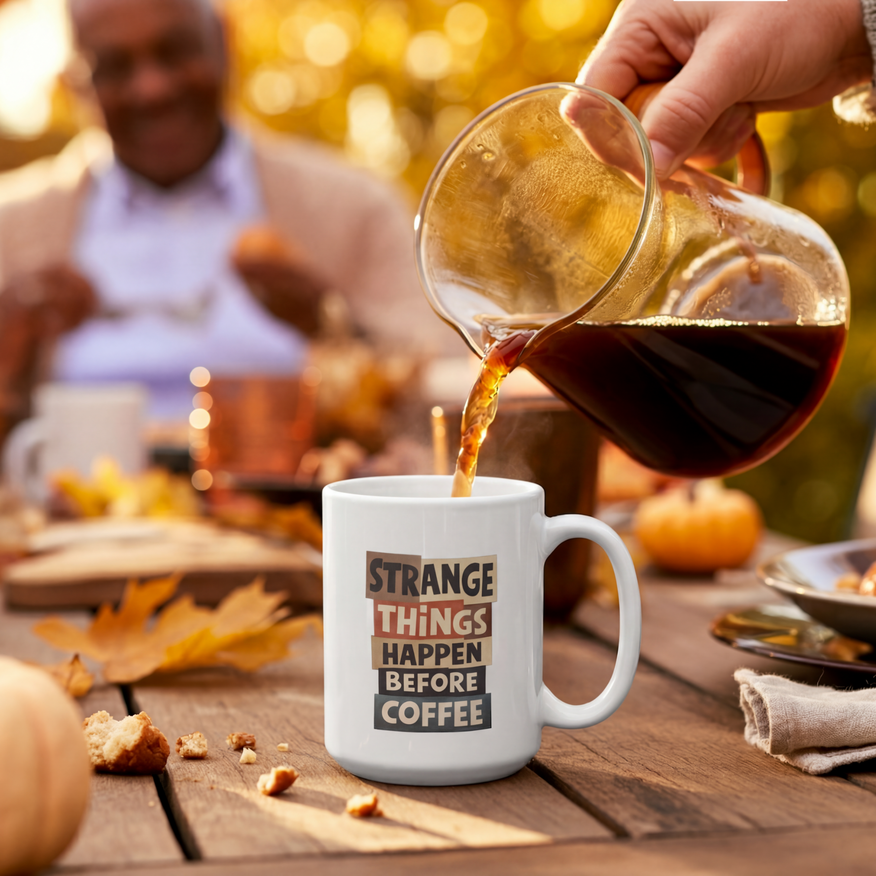 White mug with "Strange Things Happen Before Coffee" text at outdoor gathering.