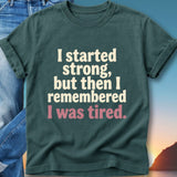 Dark green t-shirt with white and pink text reading "I started strong, but then I remembered I was tired."