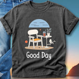 T-shirt featuring a graphic of a chair, table, drink, and bird with text "Good Day".