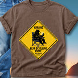Brown t-shirt with yellow warning sign: Bear sitting in chair scrolling on phone.