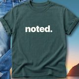 Teal t-shirt with white text "noted." on a light blue background with sky and mountains.