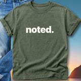 Green t-shirt with "noted." text, folded over jeans with sky background.