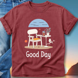 Maroon t-shirt with graphic of chair, table, drink, and bird with "Good Day" text.